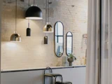 Moderne køkken med synlig murstensvæg, hængende pendler, tre ovale spejle, stablede skåle, en plante og en sort skammel ved køkkenbordet.