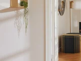 Minimalistisk indretning med en væghylde i guld med en lille plante, en sort ottoman med gulddetaljer og en gulvlampe i guld på et lyst trægulv.