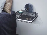En moderne væghylde i sort tråd rummer en lille grøn skål, et mørkt marmorur, smykker og en lukket notesbog ved siden af en seng med grå og beige puder.