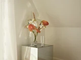 En glasvase med orange og hvide blomster og grønt står på en spejlsokkel ved siden af to tomme glas i et svagt oplyst, minimalistisk rum.