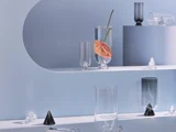 Minimalistisk hyldedisplay med klare og farvede glasvaser, hvoraf den ene indeholder en enkelt lyserød anthuriumblomst, sat op mod en lyseblå baggrund med geometriske former.