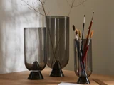 Tre røgfarvede glasvaser på en træplade; den ene indeholder fine kviste, den anden pensler, og den tredje er tom. Blødt naturligt lys skaber skygger på væggen.