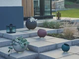 Moderne udendørs trappe med runde plantekasser med grønne planter og en sort lanterne, der fører op til en moderne bygning af glas og træ med græs og sten i baggrunden.