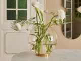 En rund glasvase med hvide blomster og grønne stilke står på et marmorbord i et blødt oplyst, moderne rum.