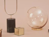 En kuglevase i glas med guldbund, en lampe i mørkt marmor og guldmetal og en lille guldkube på en beige overflade med en sart rosa baggrund.