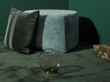 En rund grå ottoman, en stribet grå pude, en rund glasvase med guldfod og en mønstret æske på et mørkegrønt tæppe mod grønne vægge.