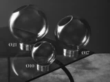 Tre glaskugler med vinklede åbninger og metalbunde, mærket Ø21, Ø17 og Ø10, er udstillet på et glasbord mod en mørk baggrund.