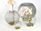 Fire runde, røgfarvede glasvaser med guldbund, tre med store kunstige blomster, arrangeret på en hvid baggrund.