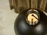 Nærbillede af en moderne lampe med en sort, mat base og en metallisk, cirkulær top med en ledning, der strækker sig fra midten. Marmorgulv og struktureret gardin i baggrunden.