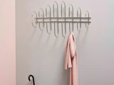 Et moderne vægstativ i metal med et abstrakt ovalt design holder et lyserødt tørklæde; en sort paraply læner sig op ad den lyse væg nedenunder.