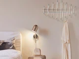 Et moderne soveværelse med en væghængt metallisk knagerække, en beige morgenkåbe, en guldlampe og et lille natbord med et ur og en bog.