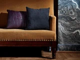 En sennepsgul polstret sofa med træben står foran en marmoreret væg. To puder, en mørklilla og en sort, hviler på sofaen.