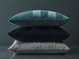 Tre rektangulære fløjlspuder i blågrønt, marineblåt og lysegråt er stablet oven på hinanden mod en mørk, dæmpet baggrund.