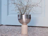 En minimalistisk plante med tynde grene står i en moderne, reflekterende vase i metal og beige på et spættet stengulv foran en lyseblå dør.