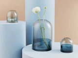 Tre blåtonede glasvaser i forskellige størrelser står på lyseblå, cylindriske sokler. Den største vase indeholder en hvid blomst med grøn stilk.