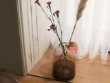 En glasvase med lyserøde nelliker, en enkelt pampasgræsstængel og en lyserød anthurium står på et trægulv ved siden af hvide gardiner.