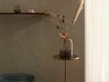 En lilla glasvase med pink og lilla blomster står på et lille rundt bord ved siden af et svagt oplyst vindue; på væggen ovenover står en hylde med dekorationer.