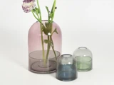 Tre glasvaser i forskellige størrelser og farver, hvor den største indeholder lilla blomster; alle har afrundede former og et minimalt design på en hvid baggrund.