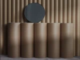 Et rundt spejl hænger på en væg med lodrette beige paneler over fem høje, cylindriske beige søjler, hvilket skaber et moderne, minimalistisk design.