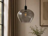 En pendel i klart glas med en pære med synlig glødetråd hænger i et blødt oplyst rum tæt på et vindue med beige gardiner og en plante i forgrunden.