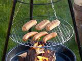 Fire pølser og tre hotdogboller grilles over åben ild på en ophængt rund metalrist med grønt græs i baggrunden.