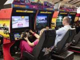 To personer spiller Daytona USA-racerspil side om side ved hver sin maskine med racerhjul og farverige skærme.