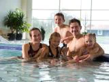 En smilende familie på fem poserer sammen i en indendørs swimmingpool, med to voksne og tre børn i vandet nær bassinkanten. Store vinduer og planter i baggrunden.