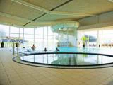 Indendørs swimmingpool med vandrutsjebane, flere personer i vandet, store vinduer, der lukker naturligt lys ind, og synlige siddepladser ved poolen.