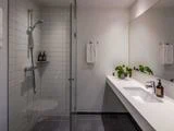 Moderne badeværelse med glasbruser, hvide flisevægge, gråt gulv, lang hvid bordplade med vask, stort spejl, potteplante og sæbedispensere.