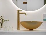 En moderne håndvask med guldarmatur og rund guldkumme ved siden af to omvendte glas, en lille plante og et håndklæde under et cirkulært spejl med baggrundsbelysning.