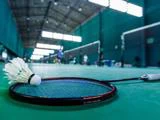En badmintonketcher og en fjerbold hviler på en grøn indendørs bane med slørede spillere og net i baggrunden.