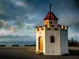 En lille hvid bygning med rødt tegltag og tårnlignende træk står alene ved havet under en dramatisk, overskyet himmel.