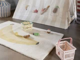 Et legerum med et bananmønstret tæppe, legetøjsmad og kurve på gulvet foran et hvidstribet telt med netlommer med legetøjsfrugter og brød.