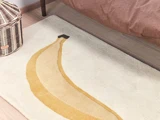 Et cremefarvet tæppe med et stort, stiliseret bananmotiv ved siden af en flettet kurv og et brunt sengetæppe på et trægulv.