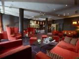 Moderne loungebar med røde lænestole og sofaer, mørke vægge, små borde med blomsterarrangementer og en bardisk med taburetter i baggrunden. Blød belysning skaber en hyggelig atmosfære.