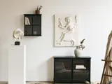 Minimalistisk rum med et sort skab, en hvid vase, en moderne abstrakt skulptur, en sort væghylde med bøger og en tekstureret vægkunst af to figurer på en hvid væg.