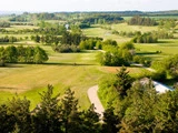 Luftfoto af en frodig grøn golfbane med snoede stier, træer og åbne fairways under en delvis overskyet himmel.