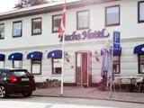 En hvid bygning med blå baldakiner mærket Techs Hotel, et dansk flag, en parkeret bil, udendørs borde og et blåt parkeringsskilt foran.