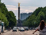 En travl gade i Berlin med biler, cyklister og træer langs vejen; Sejrssøjlen med en gylden statue står midt i baggrunden.