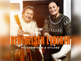 En ældre mand med en guitar og en smilende kvinde sidder sammen, begge iført strikkede trøjer. Tekst: BERTHELSEN LYBERTH og MELLEM STORM & STILHED.