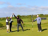 Fire personer på en golfbane; to gør high-five, mens en holder en golfkølle, og en anden holder flagstangen, alt sammen under en blå himmel med lette skyer og grønt landskab i baggrunden.