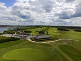 Luftfoto af en golfbane med grønne fairways, et klubhus, parkerede biler og spredte skyer på himlen. Et vandområde er synligt til venstre.