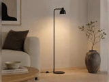 En slank, sort gulvlampe, der står mellem en sofa og en stor dekorativ vase.