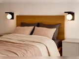 En pænt redt seng med beige og brunt sengetøj, en hovedgærde af træ, to natborde og moderne vægmonterede globelamper på hver side.