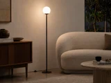 En høj, slank gulvlampe med en lysende, kugleformet pære står mellem et skab af træ og en cremefarvet, buet sofa.
