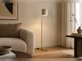 En slank gulvlampe i messing med en lille cylindrisk lampeskærm står mellem en sofa med struktur og et sidebord.