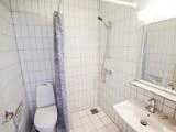 Et hvidt flisebadeværelse med toilet, håndvask, spejl, væghængt bruser og et blåmønstret bruseforhæng.