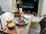 En bøfmiddag med grøntsager, pommes frites, sauce, et glas rødvin og en flaske på et træbord dækket til én person ved siden af en pejs.