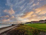 En lang bro strækker sig over vand nær en kystlinje med et græsareal og bygninger med røde tage under en farverig, overskyet himmel ved solnedgang.