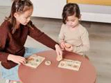 To unge piger sidder ved et rundt bord og leger med form- og billedpuslespil i træ.